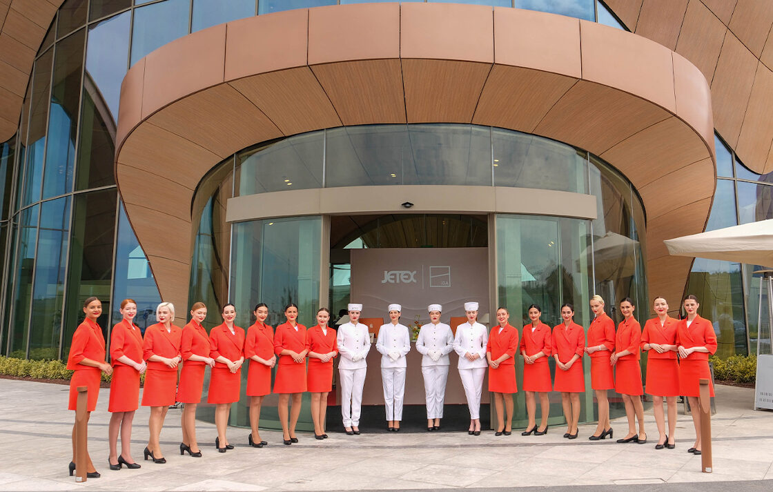 Fifteen women in red air attire with black shoes standing beside four women in all white. They're all standing in front of the new terminal building.