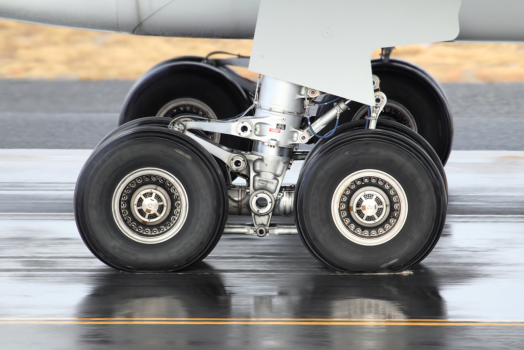 In the centre of the image the landing gear of the Airbus A330 is positioned on the ground. Four black tyres can be seen joined to the aircraft. The top of the image shows a close up of the underbelly of the aircraft. The foreground of the image is tarmac with two thin yellow lines - the aircraft is touched down at an airport. 
