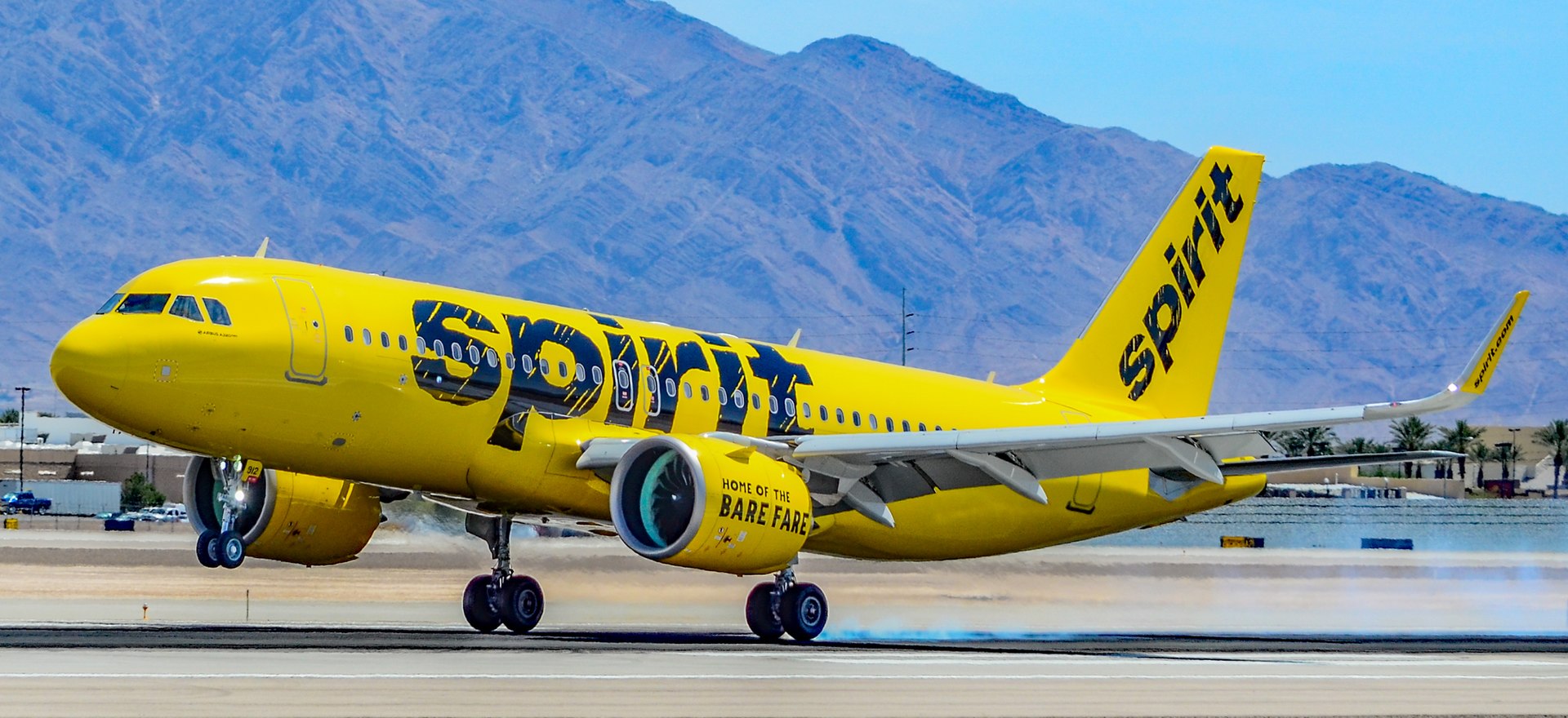 Spirit Airlines Airbus on the tarmac before takeoff. Yellow and black livery, in front of a blue sky with mountains in the background.