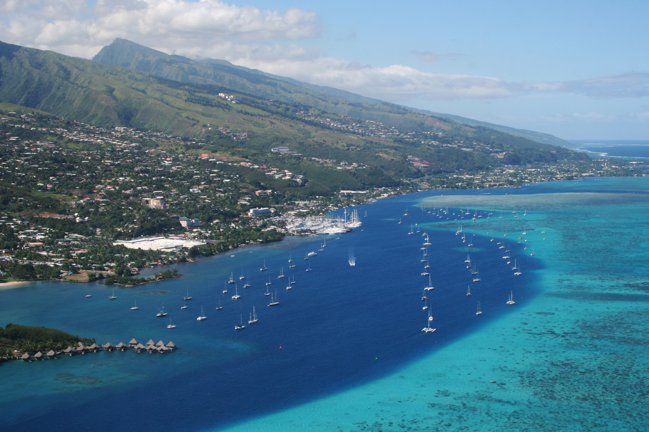 Marina Taina in Papeete. The left of the image is occupied by the shoreline and the mountains, while the right and bottom of the image shows clear blue waters. The sky in blue in the background with a few clouds above the peaks of the mountains.