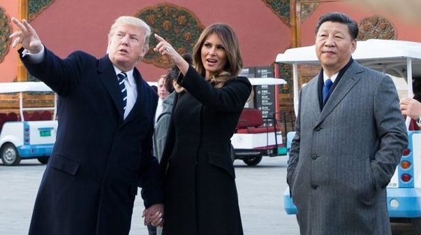 Donald Trump to the left and Melania Trump positioned to the centre left of the image, point out/up with Xi Jinping, leader of China, to the right of them looking towards what they are pointing at. The Trumps are both wearing long black trench coats, with Donald Trump wearing a white shirt and dark striped tie underneath, and Xi Jinping is wearing a grey trench coat with a blue tie, white shirt, and dark blazer collar visible at the collar. The Trumps are opening their mouths as if talking, while Xi Jinping has a pleasant neutral expression on his face. In the background a tangerine coloured wall can be seen with decorative ovals, and golf carts are scattered around them.