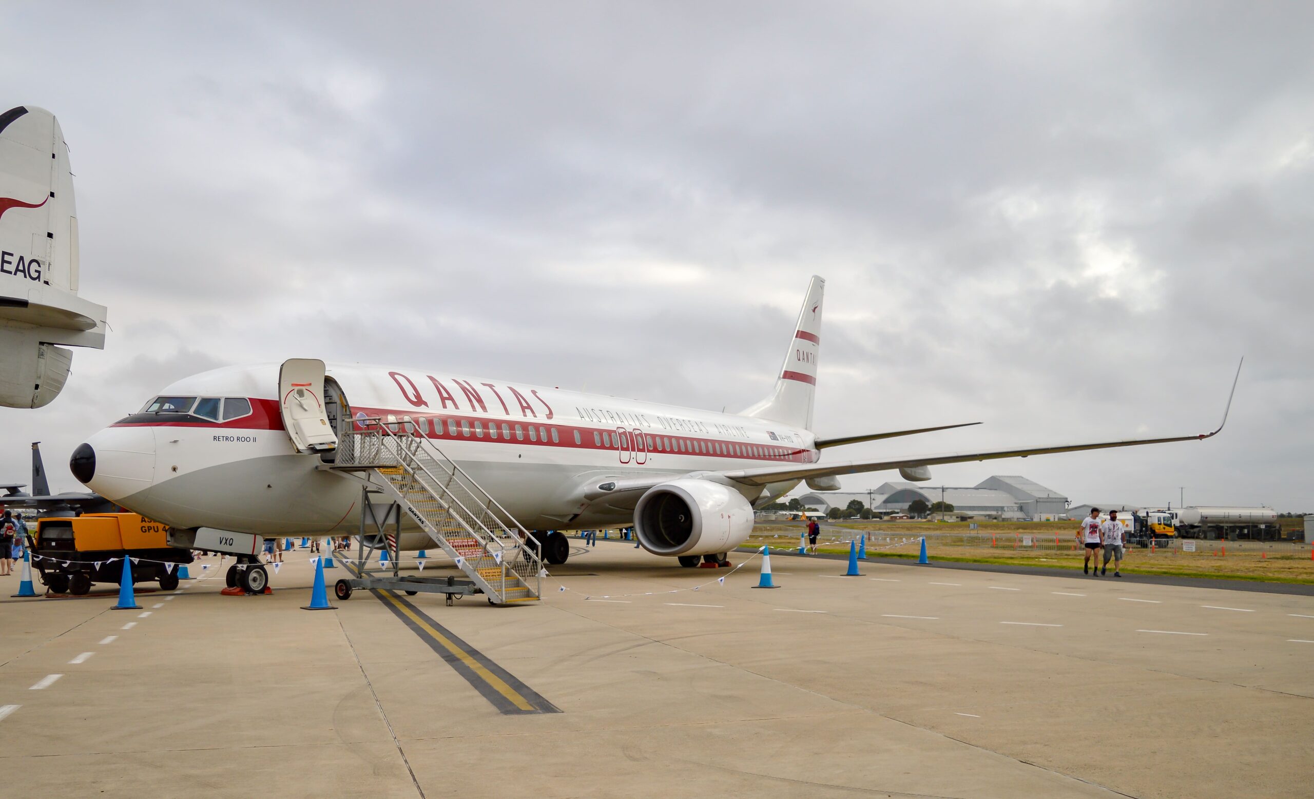 Wide view of a Qantas Boeing 737 parked on an airport apron, painted in a retro white and red livery with “QANTAS” written prominently along the fuselage and “Australian Airlines” in smaller lettering beneath the windows. The aircraft’s front door is open with a mobile stairway positioned for boarding, while a ground power unit and cables sit beneath the nose. Blue traffic cones mark a cordoned path around the aircraft, and a few people walk across the concrete apron to the right. The jet’s wing and engine extend into the frame, with low airport buildings and hangars in the distance under a heavy, overcast sky.