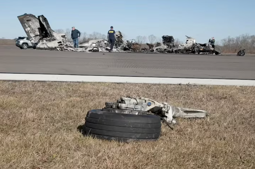 The wheel of a plane lying in the grass with the wreckage of a plane behind it