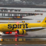A large yellow Spirit Airlines jetliner on the runway beside a body of water