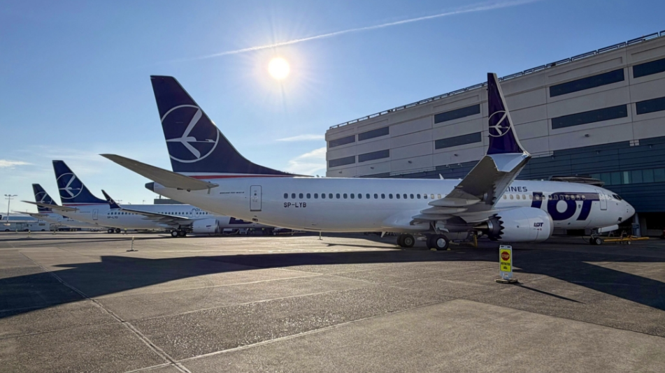A large blue and white jetliner is sitting outside an airport. Blue sky and sun in the background.
