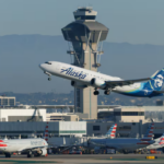 A white and blue plane taking off from a runway with an air traffic control tower