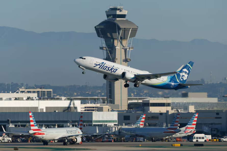 An Alaskan airlines plane taking off from a runway with an air traffic control tower in the immediate background. Other aircraft can be seen on the runway beneath.