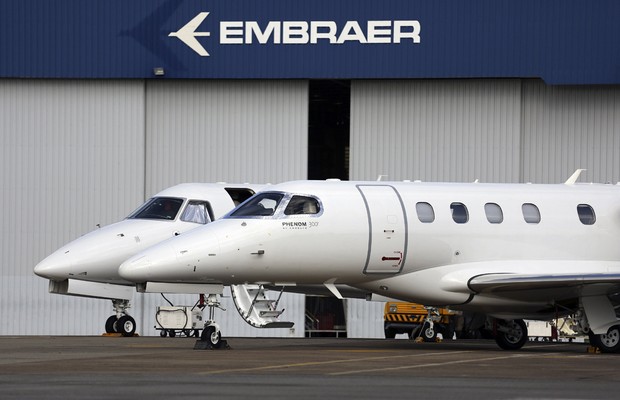 Two white Embraer business jets parked in front of an Embraer hangar building with the company logo displayed above the entrance.