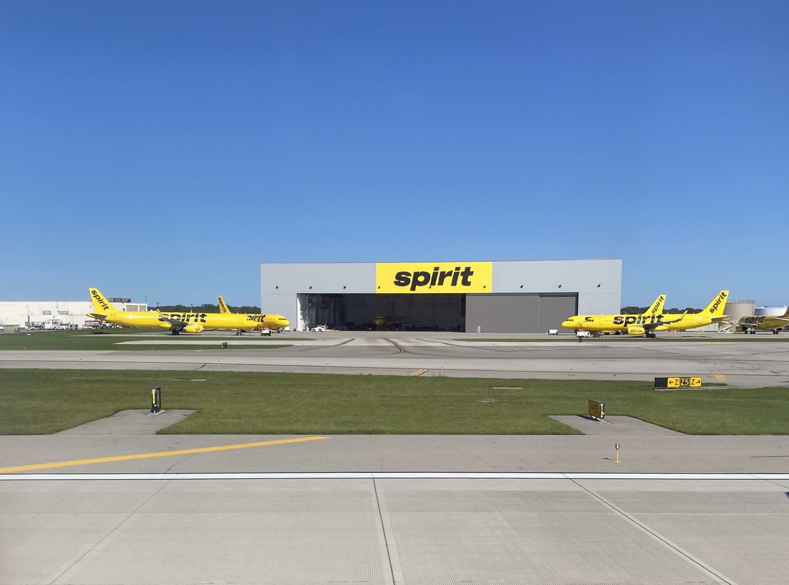 Wide view of Spirit Airlines’ maintenance area at an airport, centred on a large grey hangar with a bright yellow panel displaying the word “spirit” in black lettering above the open doors. Several yellow Spirit Airlines aircraft are parked on the apron on either side of the hangar, their tails and fuselages clearly visible against the blue sky. In the foreground, concrete taxiways and patches of green grass are marked with painted lines and signage, while the scene is brightly lit by clear daylight with no clouds overhead.
