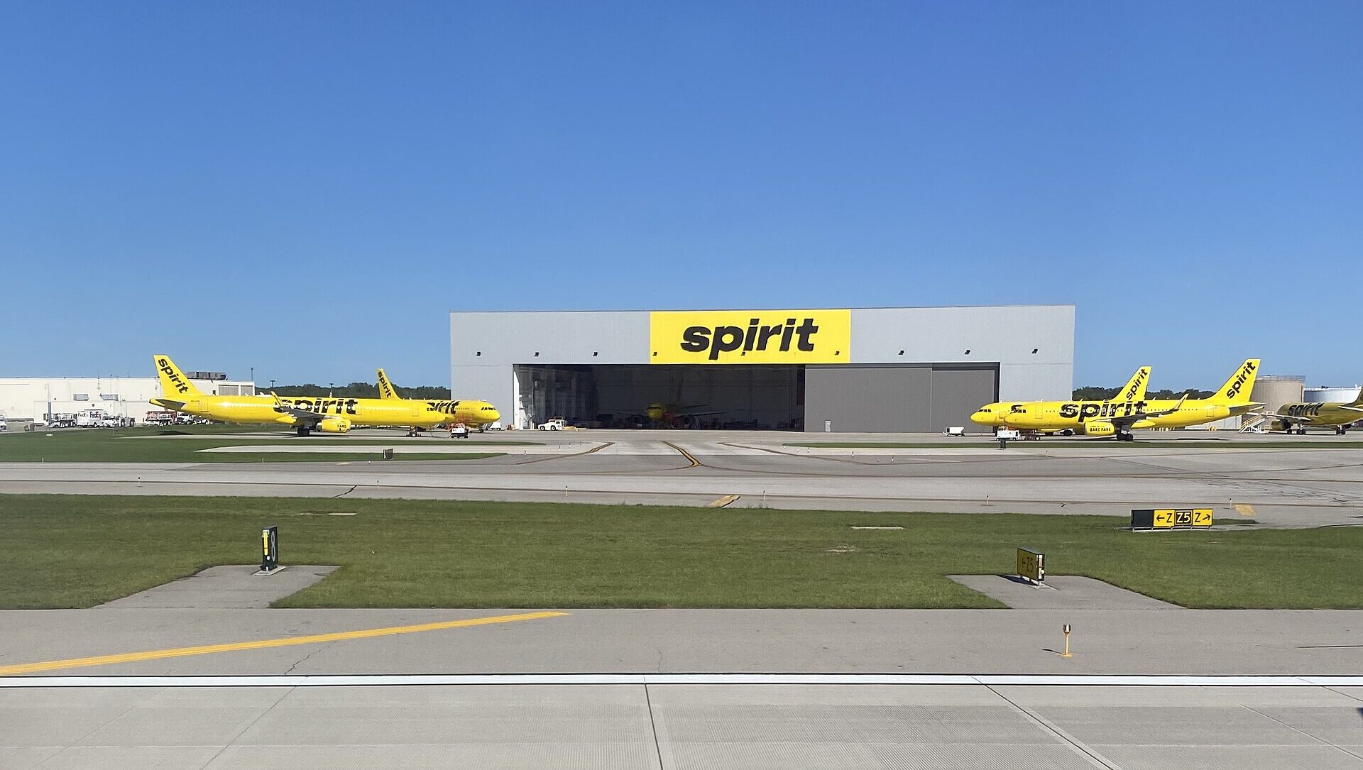 Spirit Airlines' grey building with "Spirit" written on the front in black, within a yellow box. Two aircraft in front of the building facing each other. Blue sky in the background, grass and runway in the foreground.