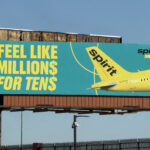 Large roadside billboard advertising Spirit Airlines against a clear blue sky. The sign has a teal background with bold yellow text reading “Feel like millions for tens,” with the dollar signs replacing the letter S. On the right side of the billboard, the bright yellow tail and rear fuselage of a Spirit Airlines aircraft are shown, with the word “spirit” printed in black on the tail and “more fly” in smaller text nearby. The billboard is mounted on a tall metal structure above a fenced roadside area, with lighting fixtures and support beams visible beneath it.