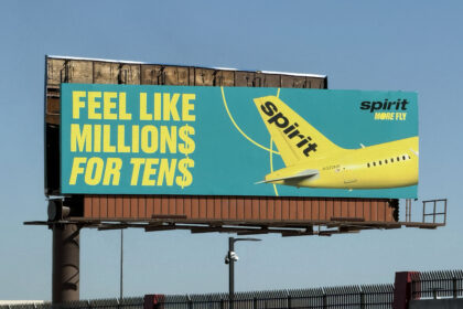 Large roadside billboard advertising Spirit Airlines against a clear blue sky. The sign has a teal background with bold yellow text reading “Feel like millions for tens,” with the dollar signs replacing the letter S. On the right side of the billboard, the bright yellow tail and rear fuselage of a Spirit Airlines aircraft are shown, with the word “spirit” printed in black on the tail and “more fly” in smaller text nearby. The billboard is mounted on a tall metal structure above a fenced roadside area, with lighting fixtures and support beams visible beneath it.