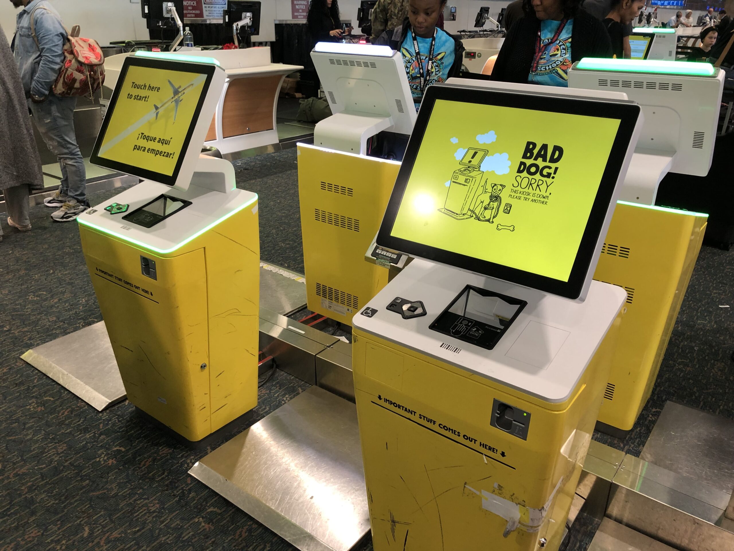 Interior view of an airport check‑in area with several yellow self‑service kiosks arranged in a row on a patterned carpeted floor. Each kiosk has a large touchscreen with green lighting along the edges; one screen displays a yellow error message reading “Bad dog! Sorry, this kiosk is down,” illustrated with a cartoon dog and machine, while another reads “Touch here to start!” in English and Spanish. The kiosks show signs of wear with scuffs and scratches on the yellow panels, and metal floor plates sit beneath them. In the background, passengers stand near counters and additional kiosks, with queue barriers and overhead signage visible under bright indoor lighting.