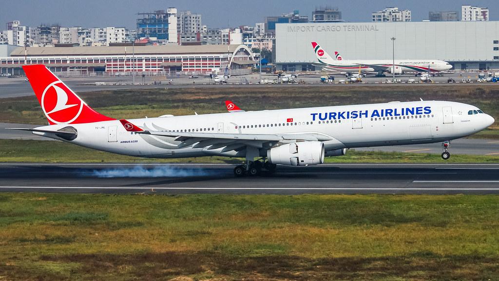 A Turkish Airlines Airbus A330 aircraft is pictured taking off on a runway. The aircraft is positioned with the nose centre right ascending and the tail centre left. Turkish Airlines branding in deep blue adorn the side of the aircraft towards the nose, and the tail is red. There is grass and tarmac in the foreground and airport buildings in the background. 