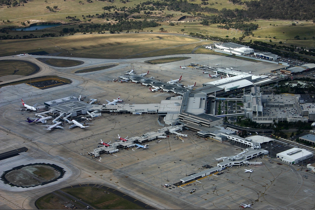 The international terminal of Tullamarine Airport in Melbourne pictured from above. Taxiways and aircraft span the majority of the image, spider webbing out from a large expanse of buildings positioned centre right of the image. There are trees upon a grassy hill in the background.