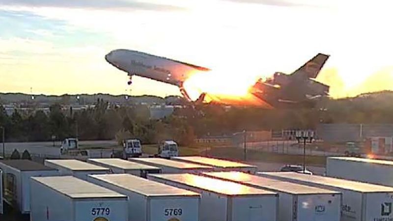 Airplane with fire visible at left pylon attachment to the wing as it clears the blast fence in footage released by NTSB.
