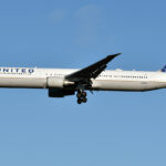 A United Airlines aircraft mid flight. the plane is white with the multi-coloured logo on the tail of the plane, which is on the centre right, and 'United' on the side of the aircraft close to the nose, which is on the centre left. The background is a clear pale blue sky.