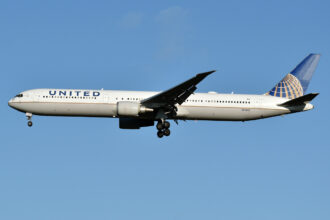 A United Airlines aircraft mid flight. the plane is white with the multi-coloured logo on the tail of the plane, which is on the centre right, and 'United' on the side of the aircraft close to the nose, which is on the centre left. The background is a clear pale blue sky.