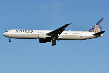 A United Airlines aircraft mid flight. the plane is white with the multi-coloured logo on the tail of the plane, which is on the centre right, and 'United' on the side of the aircraft close to the nose, which is on the centre left. The background is a clear pale blue sky.