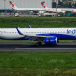 An IndiGo Airbus A321 aircraft is positioned in the centre of the image, on a tarmac runway. The nose of the plane is positioned at the centre right and the tail at the centre left. The aircraft is white with cobalt blue IndiGo branding towards the front, on the tail, and across the underbelly. The foreground and immediate background behind the plane has luscious looking green grass. The background (across the top) shows other planes a short distance away.