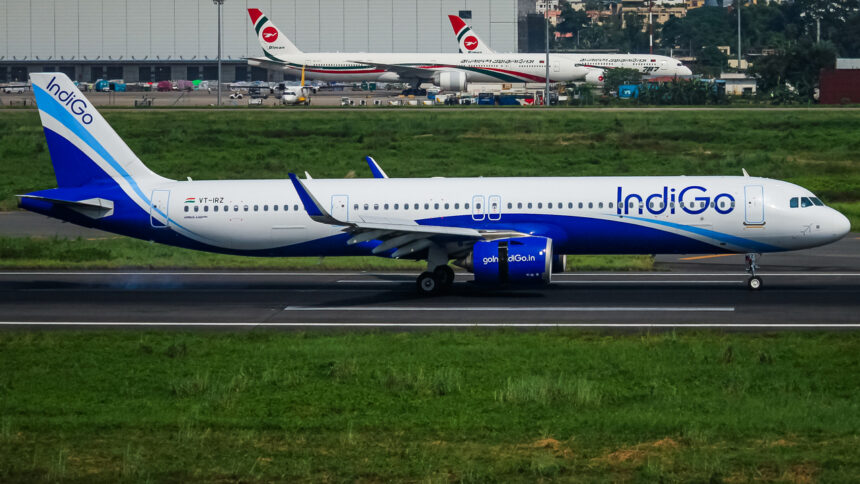 An IndiGo Airbus A321 aircraft is positioned in the centre of the image, on a tarmac runway. The nose of the plane is positioned at the centre right and the tail at the centre left. The aircraft is white with cobalt blue IndiGo branding towards the front, on the tail, and across the underbelly. The foreground and immediate background behind the plane has luscious looking green grass. The background (across the top) shows other planes a short distance away.