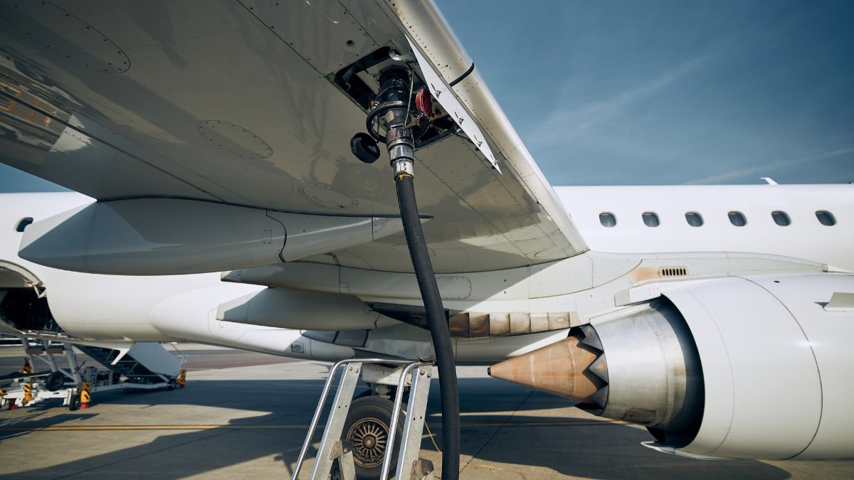 A close-up view of an Embraer E-Jet airplane being refuelled on the tarmac, showing a thick black fuel hose connected to the underside of the wing.