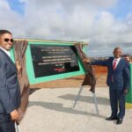 Two men dressed in suits in front of a recently unveiled plaque, celebrating the inauguration of the Negele Borana Airport. The sky is both blue and cloudy, and the ground is a mixture between sand and dirt.