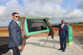Two men dressed in suits in front of a recently unveiled plaque, celebrating the inauguration of the Negele Borana Airport. The sky is both blue and cloudy, and the ground is a mixture between sand and dirt.