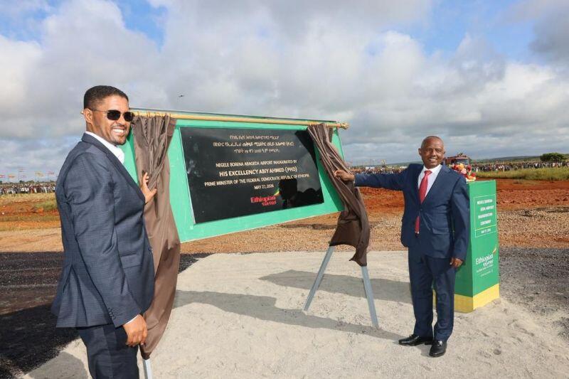 Two men dressed in suits in front of a recently unveiled plaque, celebrating the inauguration of the Negele Borana Airport. The sky is both blue and cloudy, and the ground is a mixture between sand and dirt.
