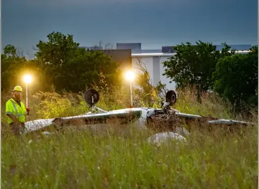 The plane crash site in Texas. Plane is upside down with the wheels visible. A man in high visibility clothing and hard hat stood on the left. Trees and buildings in the background.