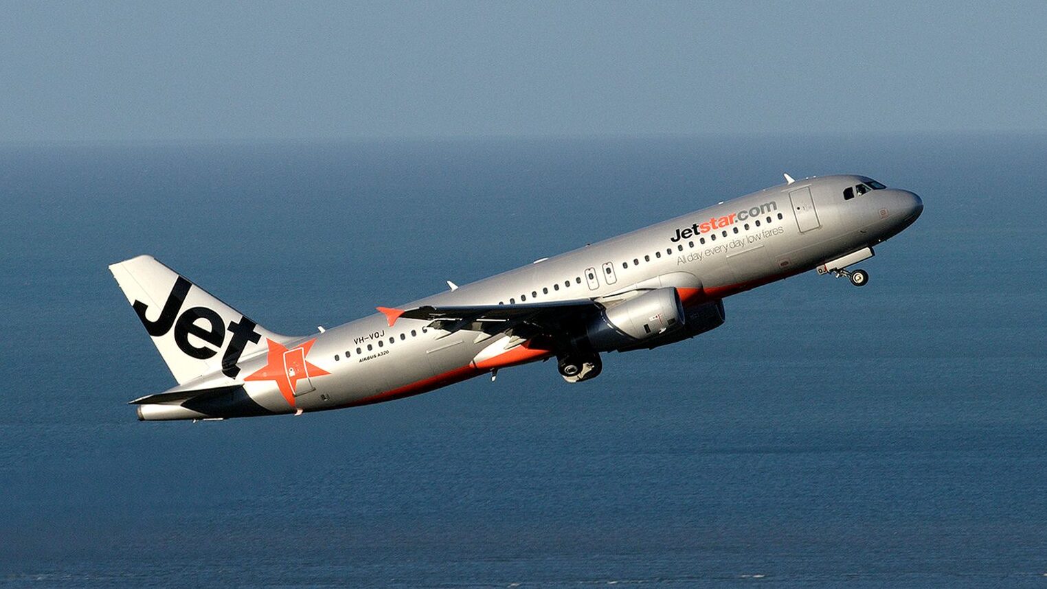 Jetstar Airbus A320 in flight over the ocean. Grey-ish sky in the background. Grey, orange and black livery.