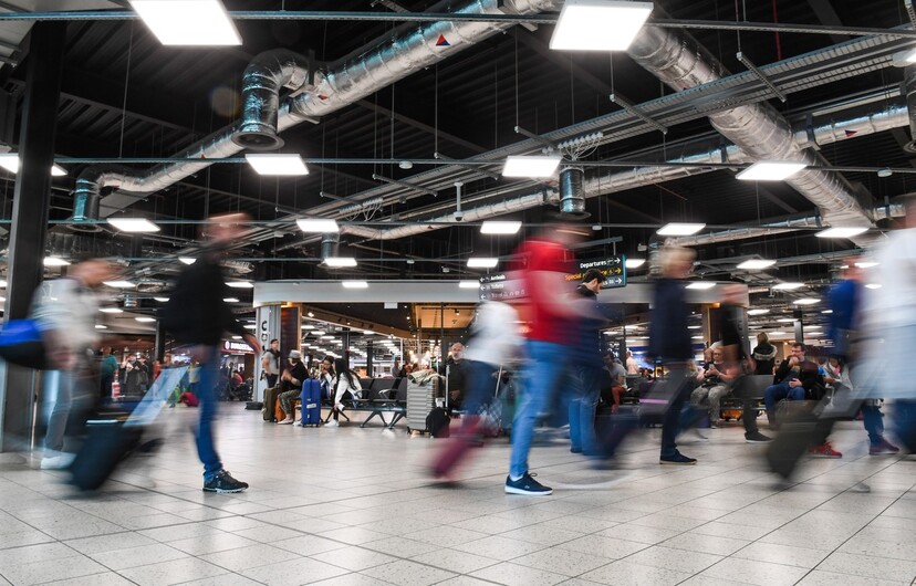 Passengers walking through LLA departures terminal, people are blurred, holding suitcases, shops in the background.