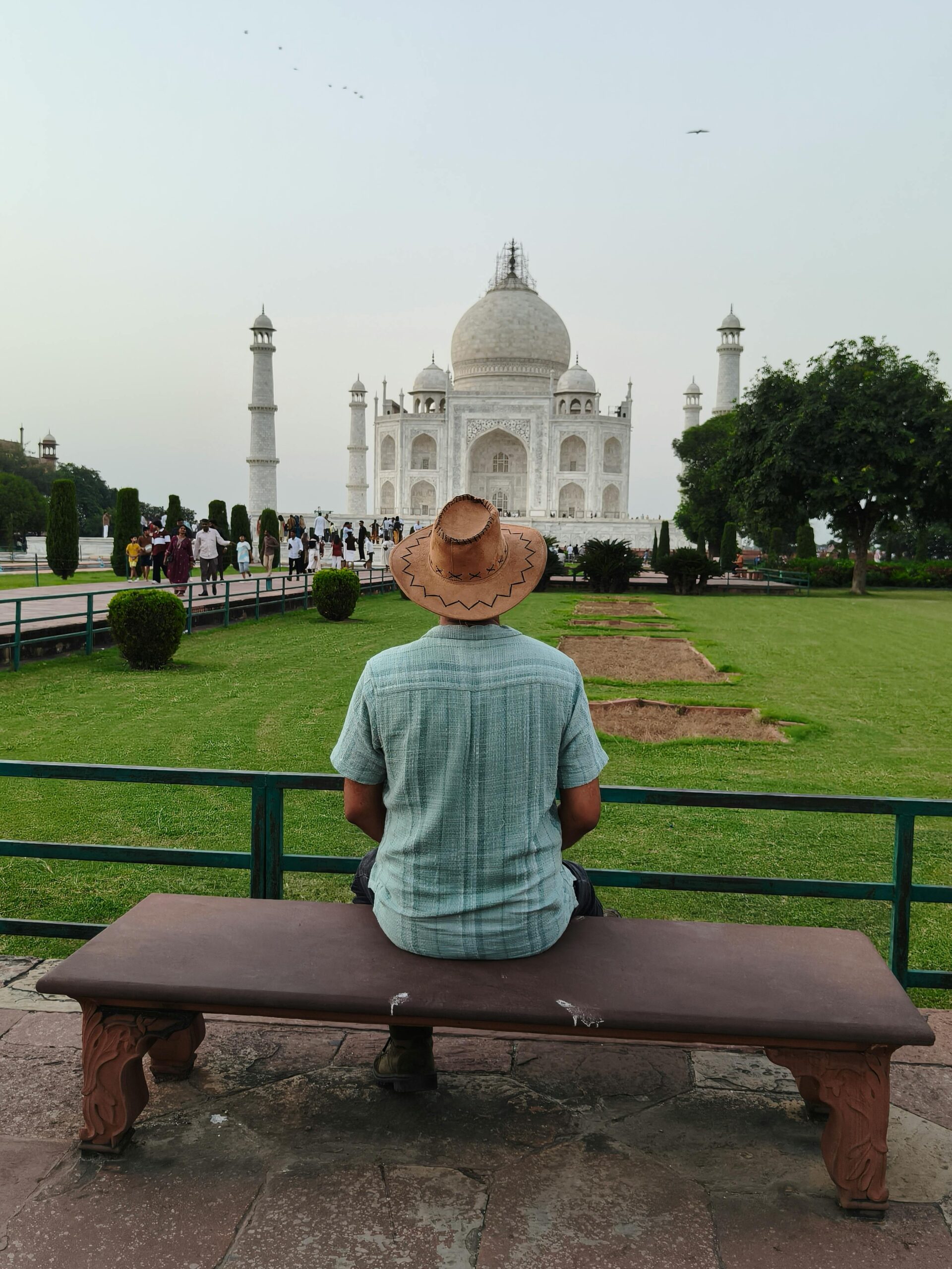 A man wearing a cowboy hat sits on a bench, observing the iconic Taj Mahal in Agra, India.