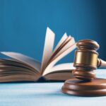 Generic image of a brown wooden court gavel and an open book behind it. The two object are placed on a white table surface. The background is a mid turquoise blue colour. The whole image is slightly stylistically blurred.