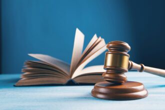 Generic image of a brown wooden court gavel and an open book behind it. The two object are placed on a white table surface. The background is a mid turquoise blue colour. The whole image is slightly stylistically blurred.