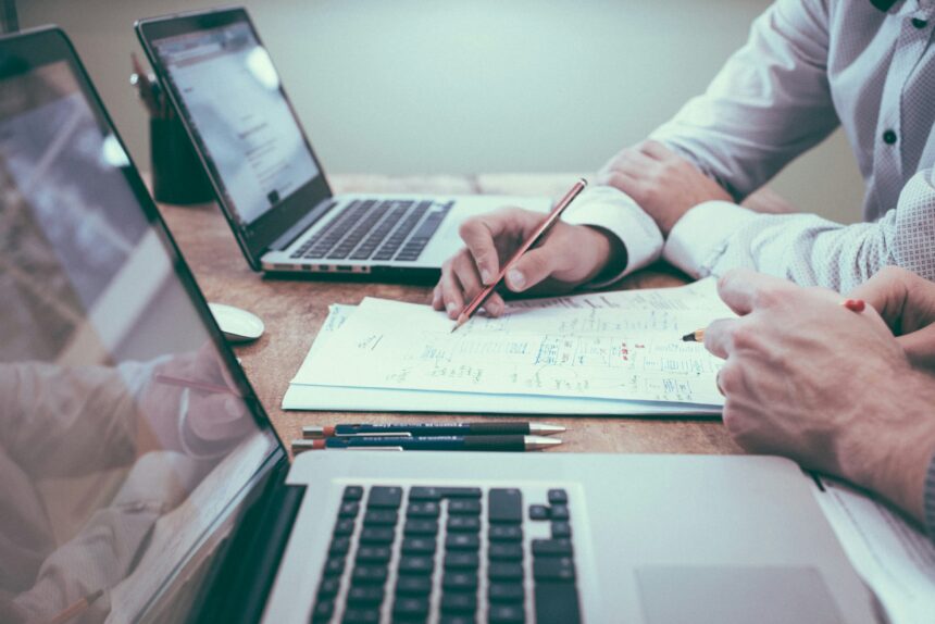Generic image of a desk with two laptops upon it and some paper. The image is taken from low down on the desk very close to one laptop. The background is stylistically blurred, but the hands and forearms can be se of two people sat at the long desk.
