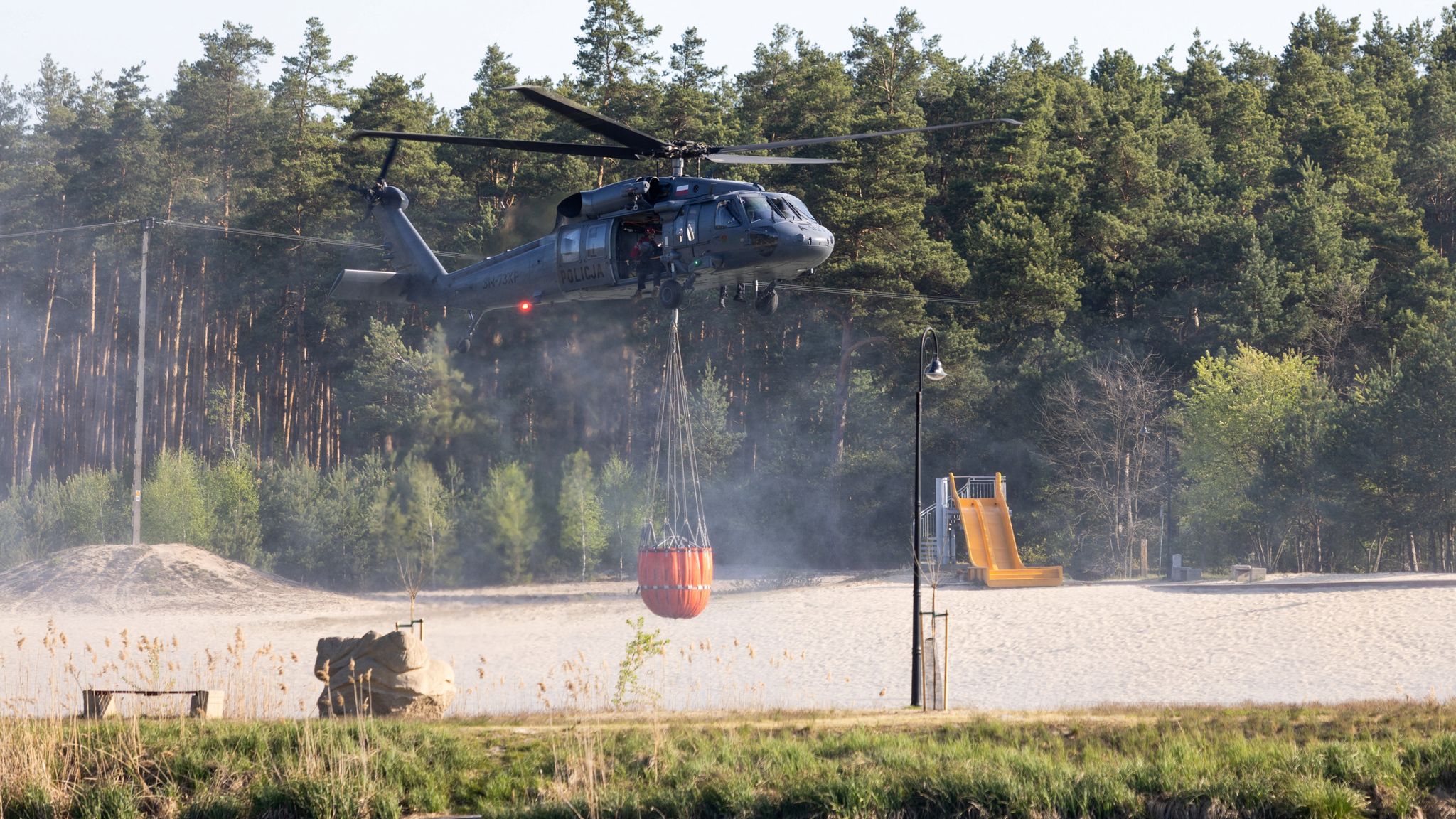 A black helicopter carrying an orange bucket of water. Forest in the background. Smoke all around.