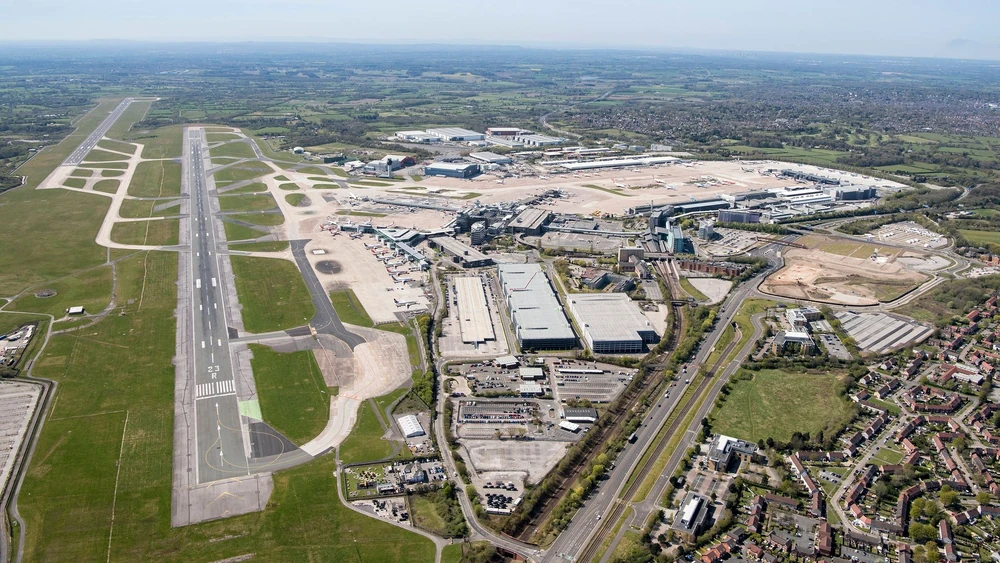 Aerial shot of Manchester Airport. Runway in view on the left side, buildings on the right. Housing estates on the bottom right behind the airport, grass all around.