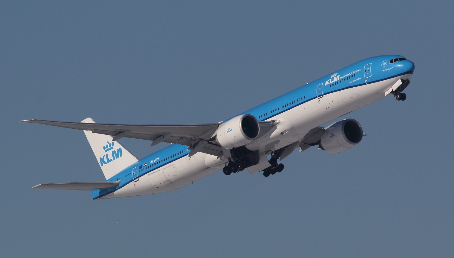 The KLM Boeing 777-300ER aircraft flying upwards toward the top right, landing gear is being tucked up. Blue/grey sky in the background.