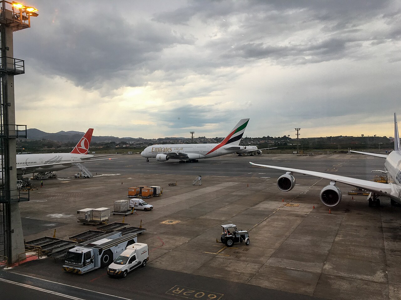 The view from inside of São Paulo's international terminal. There are three parked aircraft and a few parked vehicles. The sky is both cloudy and blue. 