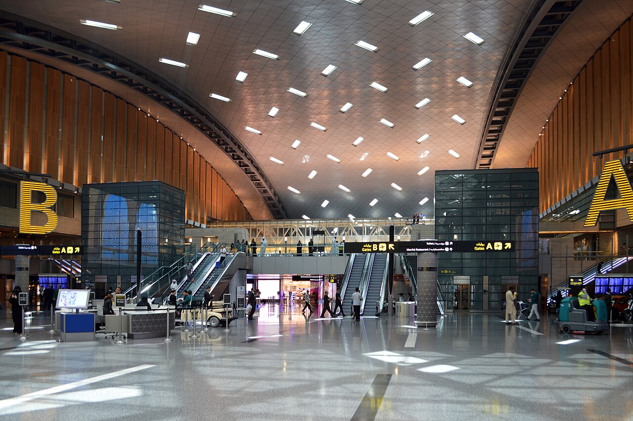 Interior view of a modern airport terminal at Hamad international airport, with a high curved ceiling, illuminated ceiling panels, escalators, and passengers walking through a spacious concourse with gate signage.