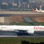 An Ethiopian Airlines Boeing 777 wide-body jet taxiing on a runway, shown in profile view. The white aircraft features the airline's name in large red lettering along the fuselage and the distinctive green, yellow, and red tail livery. Near the front, Amharic script and the name "Ras Dashen" appear above the windows, with registration "ET-ASK" visible toward the rear. In the background, a dense urban cityscape with high-rise buildings rises behind the airport, while other parked aircraft are visible on distant taxiways under a hazy sky.