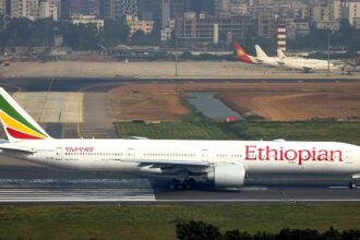 An Ethiopian Airlines Boeing 777 wide-body jet taxiing on a runway, shown in profile view. The white aircraft features the airline's name in large red lettering along the fuselage and the distinctive green, yellow, and red tail livery. Near the front, Amharic script and the name "Ras Dashen" appear above the windows, with registration "ET-ASK" visible toward the rear. In the background, a dense urban cityscape with high-rise buildings rises behind the airport, while other parked aircraft are visible on distant taxiways under a hazy sky.