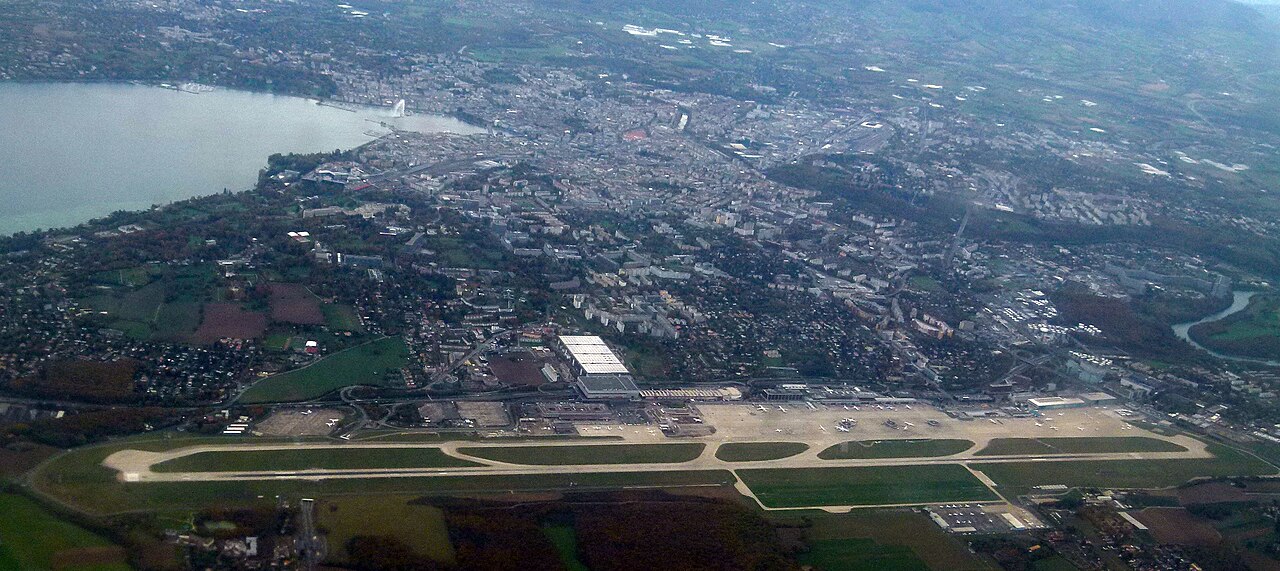 Aerial view of Geneva Airport in Switzerland showing the runway, terminal facilities, surrounding roads, and urban areas near the French-Swiss border.