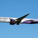 A Riyadh Air B787-9 Dreamliner flying under a blue sky. The plane's livery is mostly white with purple and lilac accents.