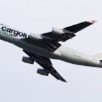 Cargolux Italia Boeing 747 freighter in flight during climb-out, captured against a pale overcast sky. The aircraft is shown banking slightly upward with landing gear still extended, highlighting the aircraft shortly after takeoff.