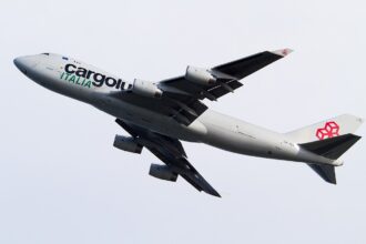 Cargolux Italia Boeing 747 freighter in flight during climb-out, captured against a pale overcast sky. The aircraft is shown banking slightly upward with landing gear still extended, highlighting the aircraft shortly after takeoff.