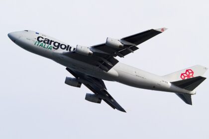 Cargolux Italia Boeing 747 freighter in flight during climb-out, captured against a pale overcast sky. The aircraft is shown banking slightly upward with landing gear still extended, highlighting the aircraft shortly after takeoff.