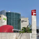 Outside view of Lisbon Airport's terminal one. The building is a mixture of light grey with a big glass wall. The sky is blue and red tube sines can also be seen