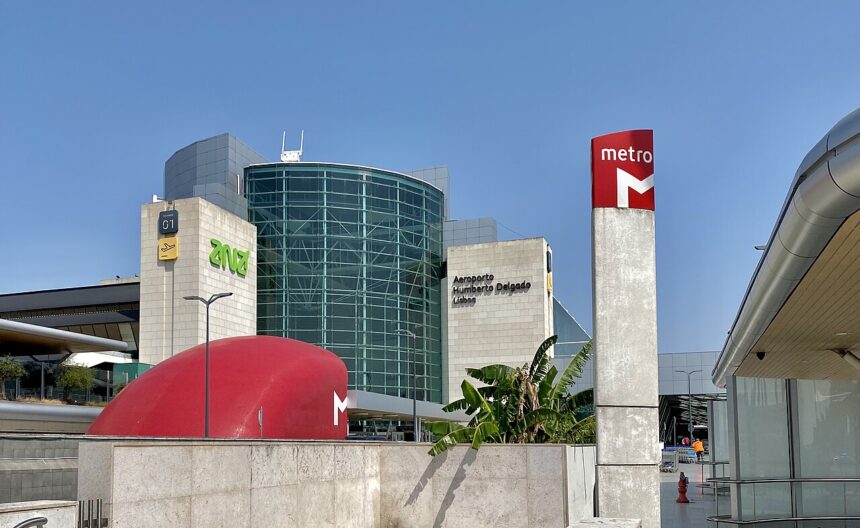 Outside view of Lisbon Airport's terminal one. The building is a mixture of light grey with a big glass wall. The sky is blue and red tube sines can also be seen